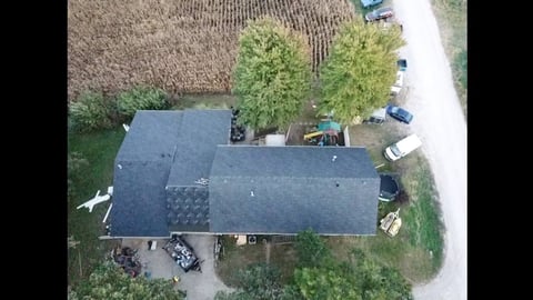 Aerial view of a single-story house with dark roof, driveway, and surrounding trees and farmland