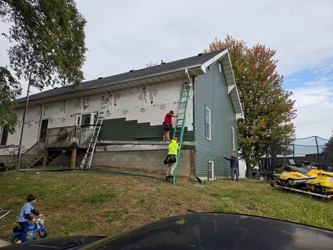 Workers in safety gear installing new siding on a two-story house under construction with ladders and equipment visible