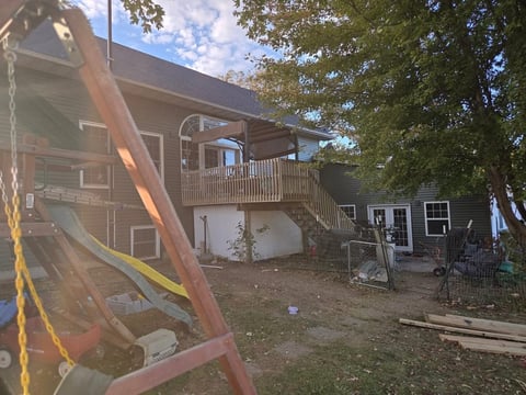 Brown wooden house with elevated deck and attached garage, surrounded by trees and construction materials in yard