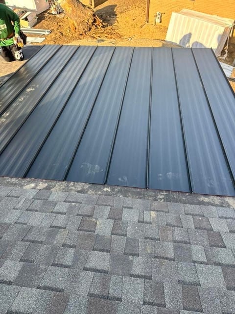 Gray corrugated metal roofing panels laid on asphalt shingles with construction materials and dirt in background