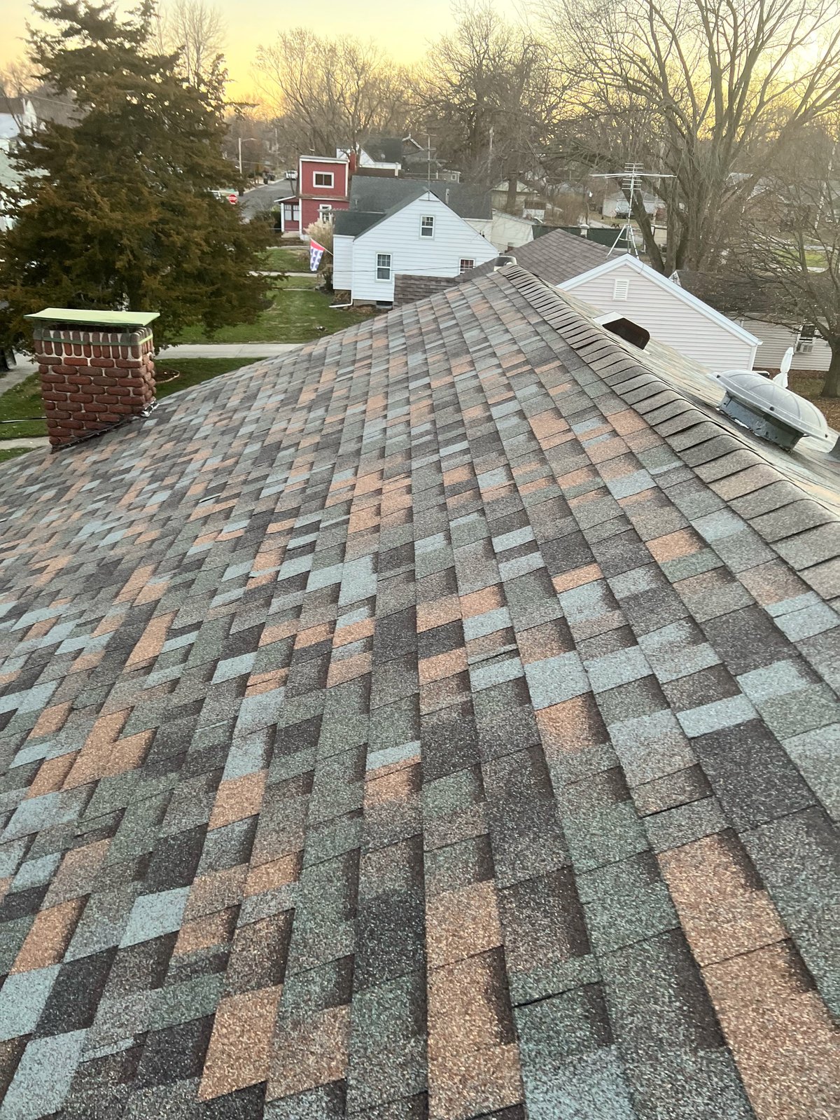 Aerial view of a residential roof with multi-colored asphalt shingles, overlooking neighboring houses and bare trees at dusk