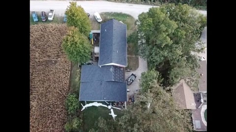 Aerial view of a single-story house with dark roof surrounded by mature trees and adjacent lawn