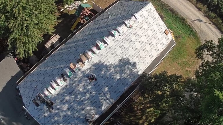 Aerial view of a large commercial warehouse roof with white panels and multiple vents, surrounded by green trees and roads