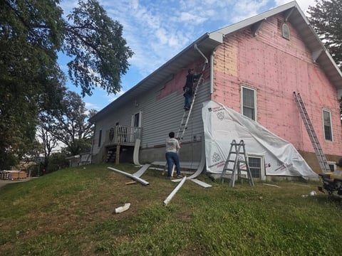 Home renovation in progress with pink brick exterior, white tarp, ladder, and construction equipment on lawn