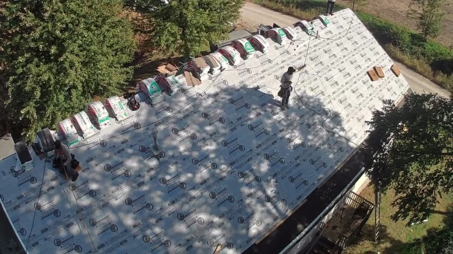 Aerial view of a house roof with white underlayment, workers installing shingles, and surrounding trees and landscaping